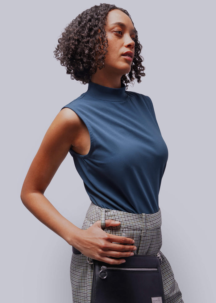 S=Woman wearing a blue sleeveless top and plaid pants, holding a black bag against a gray background