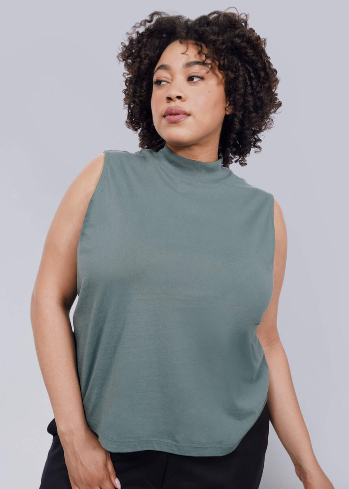 1X= A woman with curly hair models The Essential Mock Neck Tank in sage green, paired with black pants. She stands against a white background, looking calm—this relaxed-fit tank is perfect for layering with any outfit.