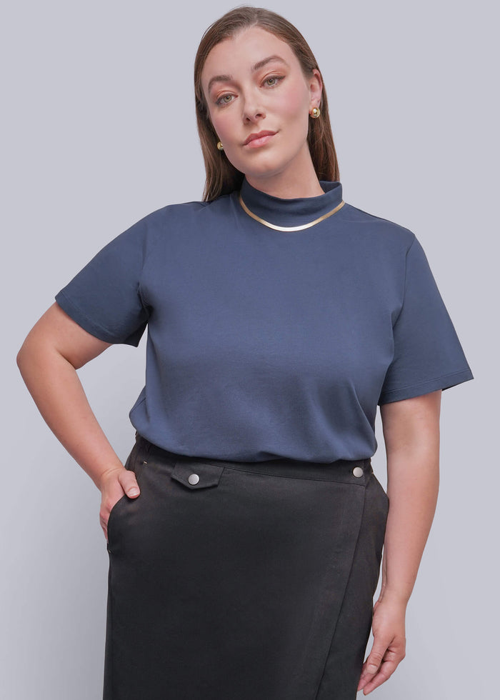 1X=A woman modeling a slate blue short-sleeve mock neck top paired with a black wrap skirt, set against a solid light gray background.