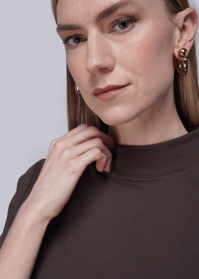 S=Close-up of a person wearing brown shirt and gold earrings against a light gray background