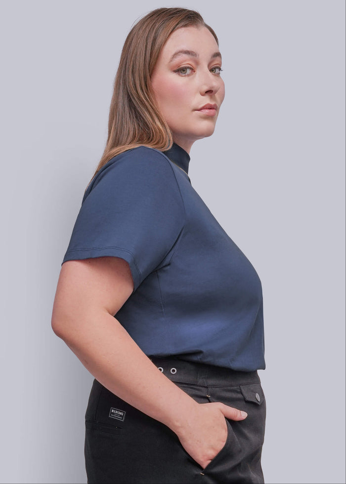 1X=A woman in a side profile wearing a navy blue short-sleeve mock neck top and black trousers, photographed against a solid light gray background.