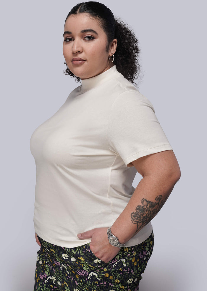 1X=Woman wearing a white mock neck t shirt and floral pants against a plain background