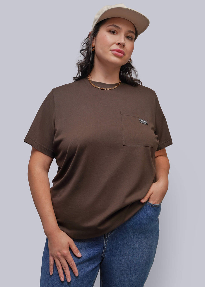 1X=A woman modeling a brown short-sleeve crewneck t-shirt with a chest pocket, paired with blue jeans and a beige baseball cap, against a solid gray background.