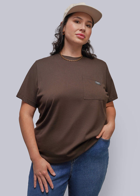 1X=A woman modeling a brown short-sleeve crewneck t-shirt with a chest pocket, paired with blue jeans and a beige baseball cap, against a solid gray background.