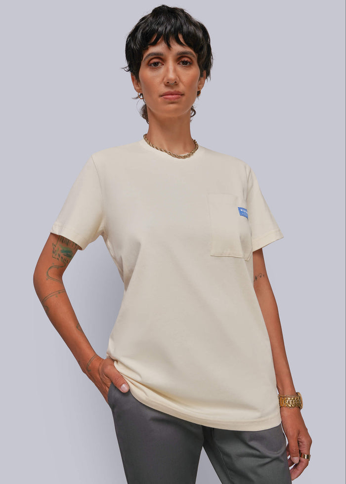 S=A model wearing a cream-colored short-sleeve pocket t-shirt with a small blue logo on the chest pocket, paired with gray pants, against a solid light gray background.