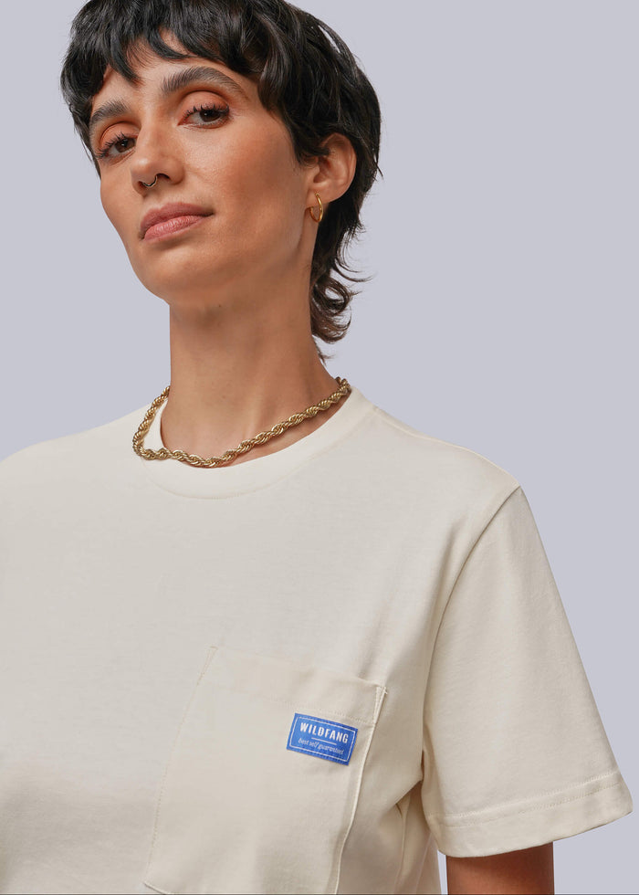 S=A close-up of a model wearing a cream-colored short-sleeve pocket t-shirt with a blue 'MIRIAM' brand label on the chest pocket, set against a solid light gray background.