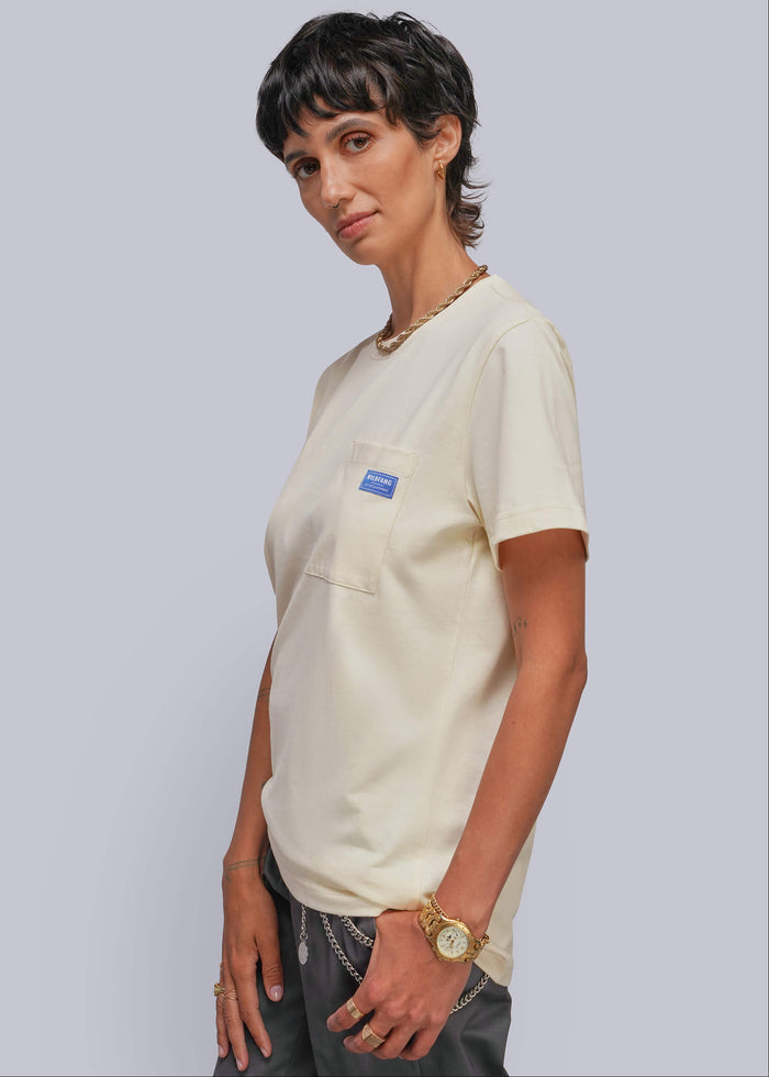 S=A person modeling a cream-colored short-sleeve pocket t-shirt with a small blue rectangular patch on the chest pocket, paired with dark gray pants, against a solid light gray background.