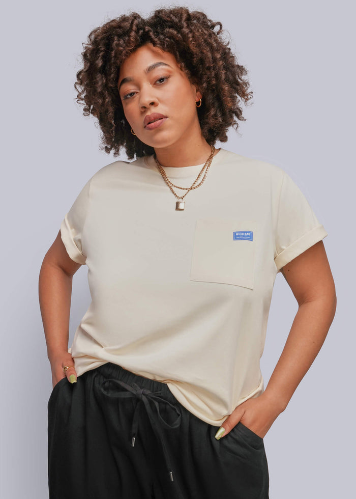 1X=A woman with curly brown hair wearing a cream-colored short-sleeve pocket t-shirt and black drawstring pants, standing against a solid light gray background.