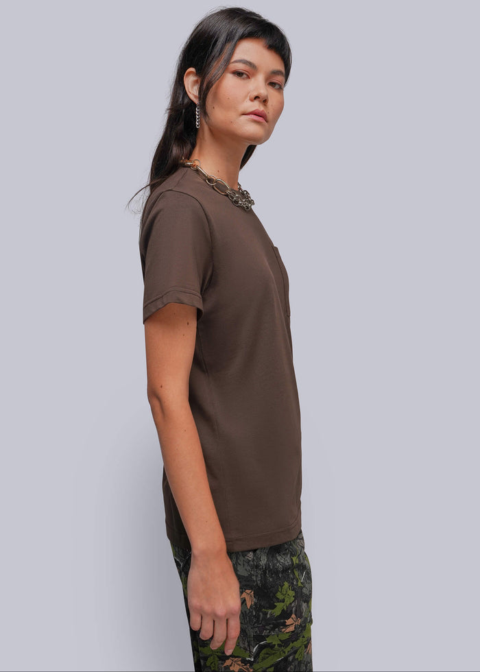 S=A woman wearing a brown short-sleeve crewneck t-shirt and a dark green floral print skirt, standing against a solid light gray background.