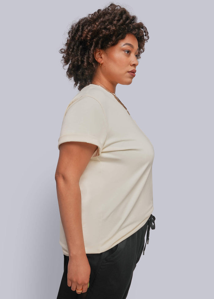 1X=A woman with curly dark hair is shown in profile wearing a cream-colored short-sleeve t-shirt and black pants against a solid light gray background.