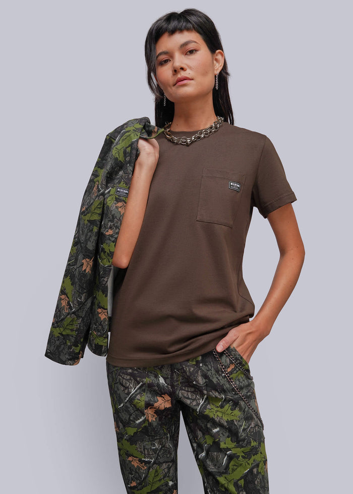 S=A model wearing a brown short-sleeve pocket t-shirt and matching camouflage pants, with a camouflage jacket draped over her shoulder, against a solid gray background.