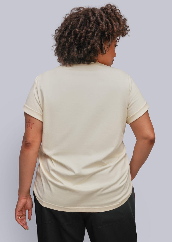 1X=A person shown from the back wearing a cream-colored short-sleeve t-shirt and black pants, standing against a solid light gray studio background.