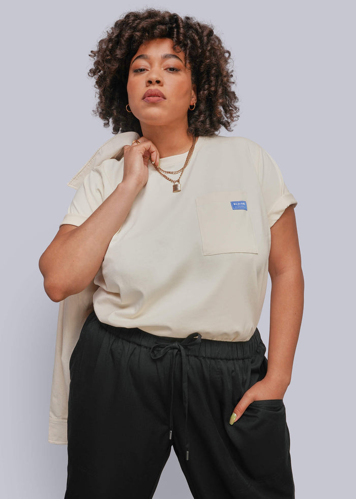 1X=A woman modeling a cream-colored short-sleeve pocket t-shirt with a small blue logo patch on the chest, paired with black drawstring sweatpants, against a solid light gray background.