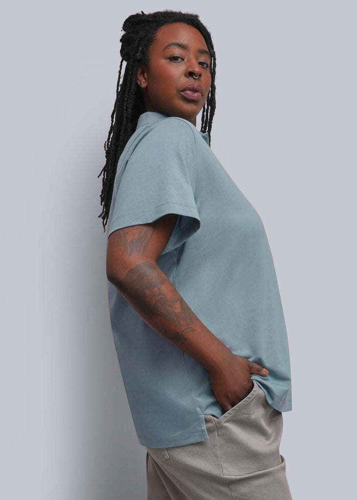 1X=A woman with long black dreadlocks is shown from a side-back angle wearing a light dusty blue short-sleeve t-shirt and beige trousers against a plain light gray studio background.