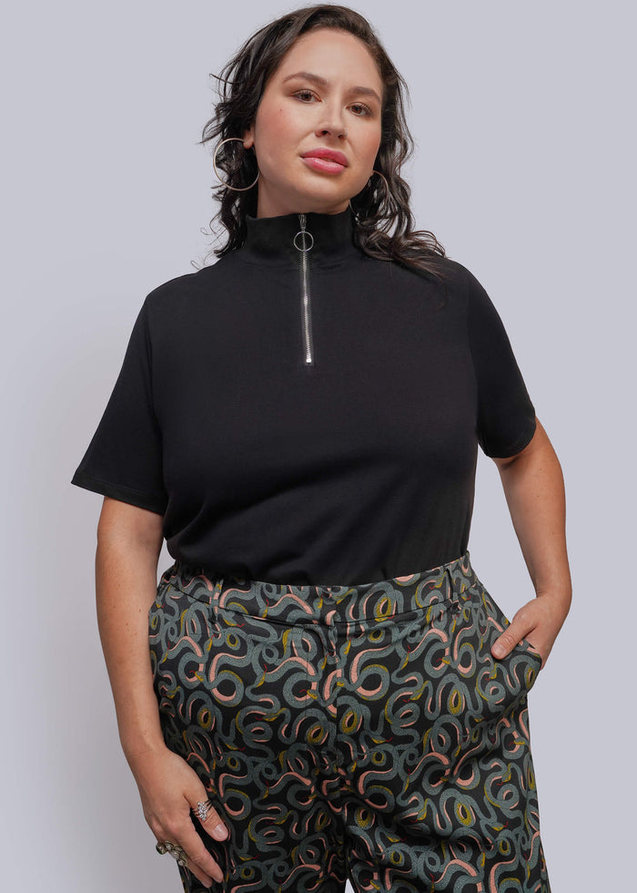 1X=A woman modeling a black short-sleeve mock neck top with a front zipper, paired with patterned green pants, against a solid gray studio background.