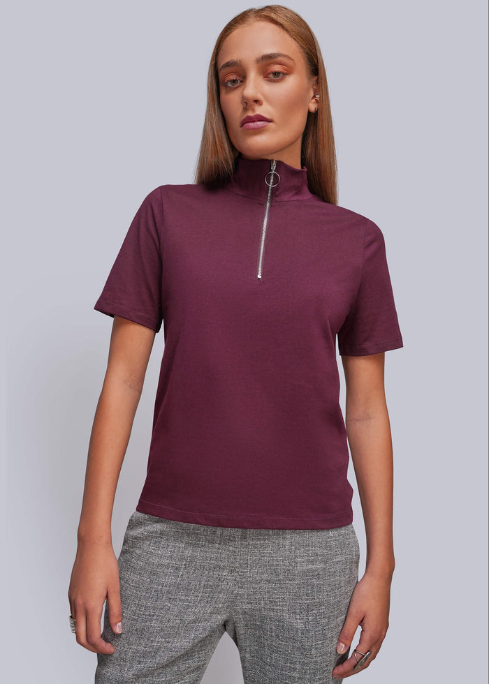 S=A woman modeling a short-sleeve mock neck top with a silver zipper at the collar, paired with gray textured pants, against a solid light gray background.
