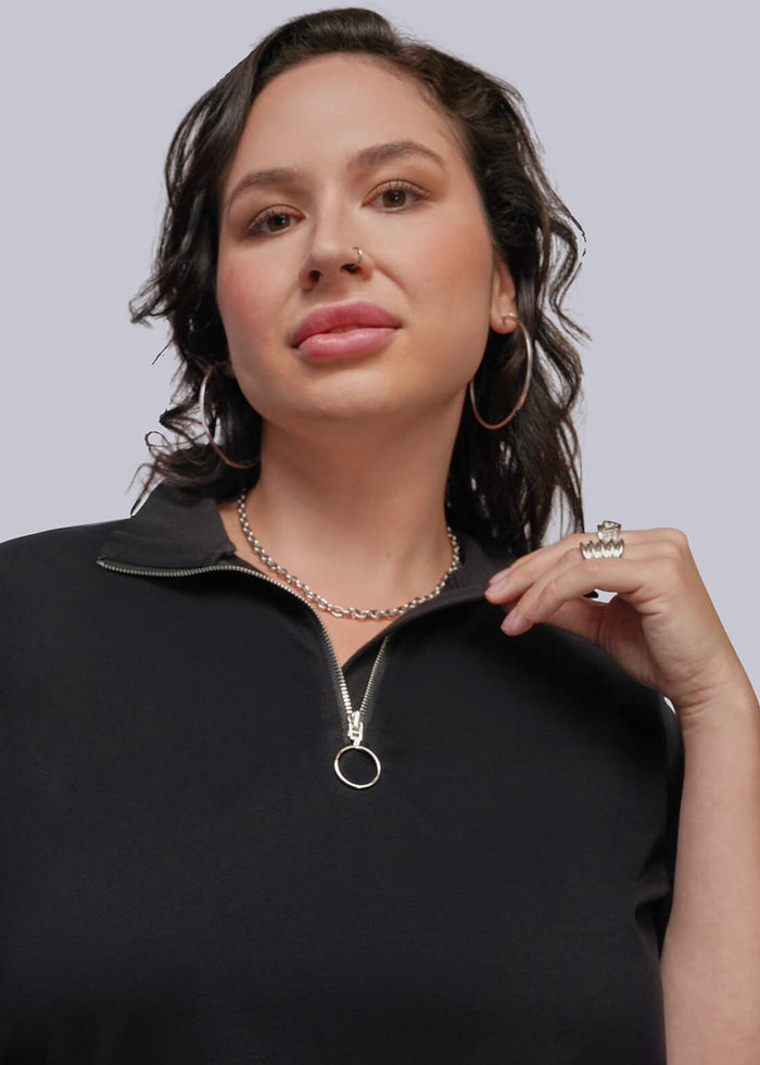 1X=A woman modeling a black short-sleeve quarter-zip polo shirt with a silver ring detail on the collar, paired with patterned pants, against a solid gray background.