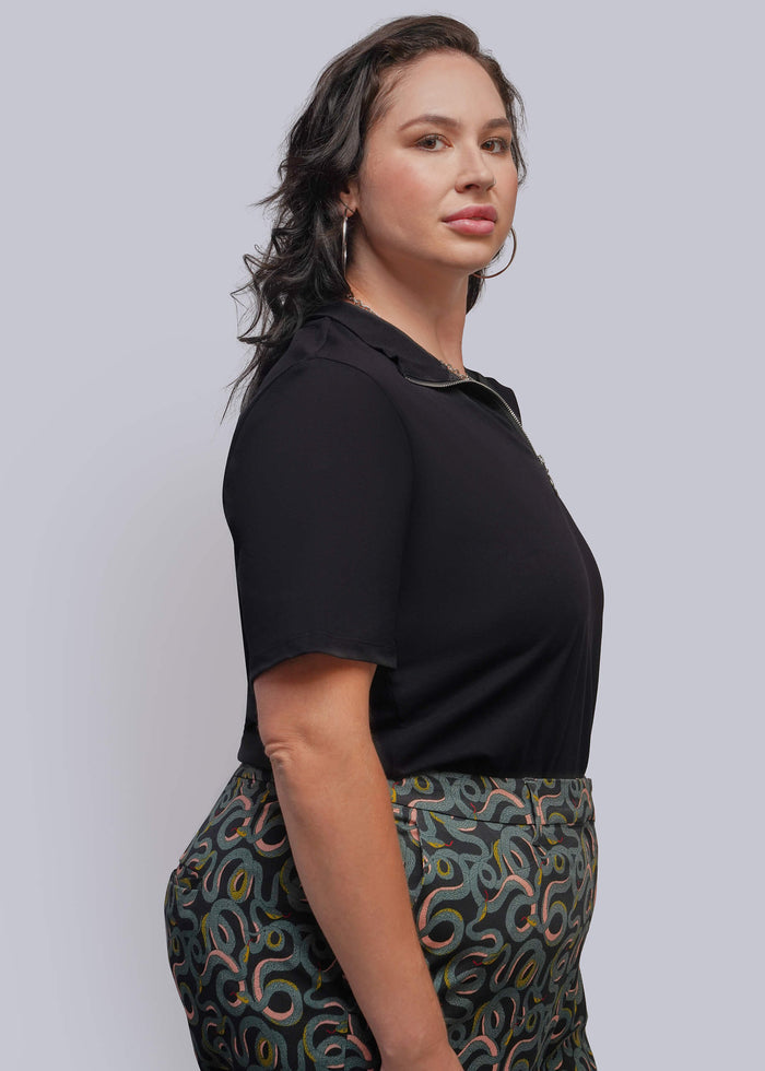 1X=A woman shown in side profile wearing a black short-sleeve polo shirt and patterned high-waisted trousers, against a solid light gray studio background.
