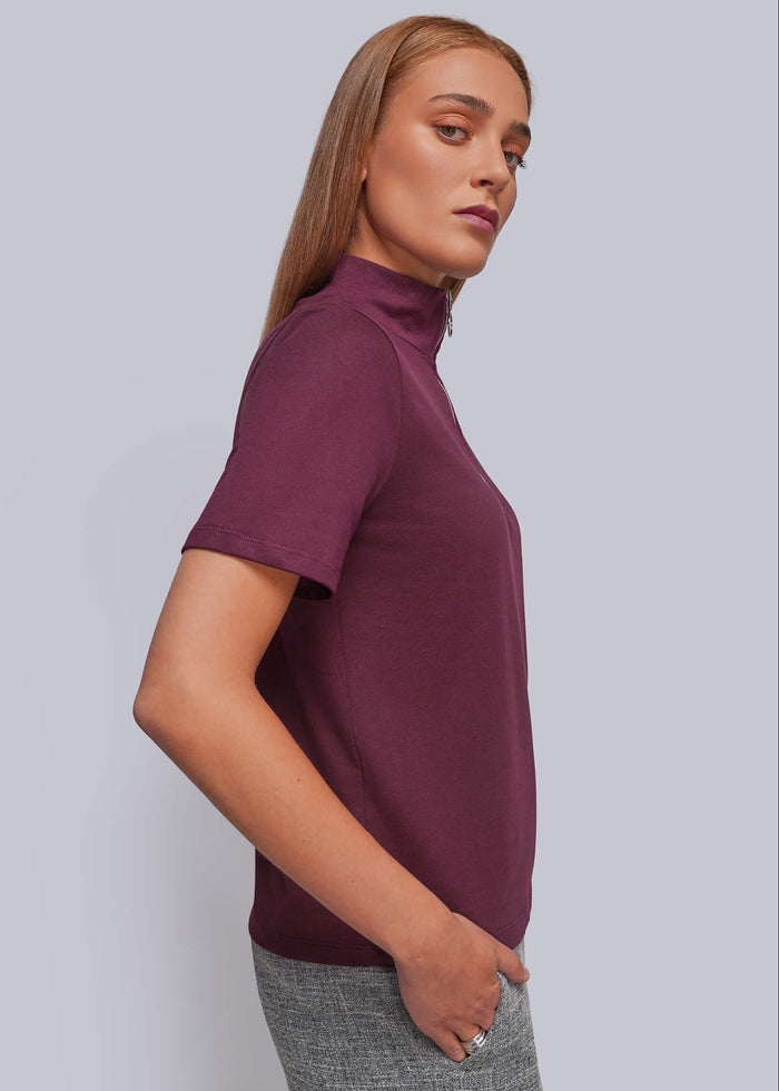 S=A woman wearing a short-sleeve mock neck top in deep burgundy, paired with gray textured pants, photographed against a solid light gray studio background.