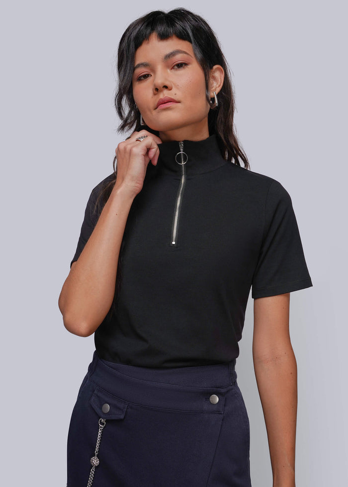 S=A woman modeling a black short-sleeve mock neck top with a silver zipper detail at the collar, paired with dark navy denim wrap skirt, against a solid gray studio background.
