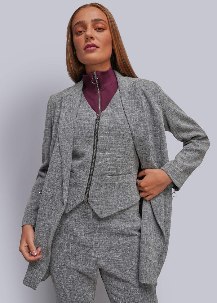 S=A woman modeling a gray three-piece suit with a textured tweed pattern, featuring a jacket, vest, and matching trousers, layered over a burgundy mock-neck top, against a solid gray studio background.