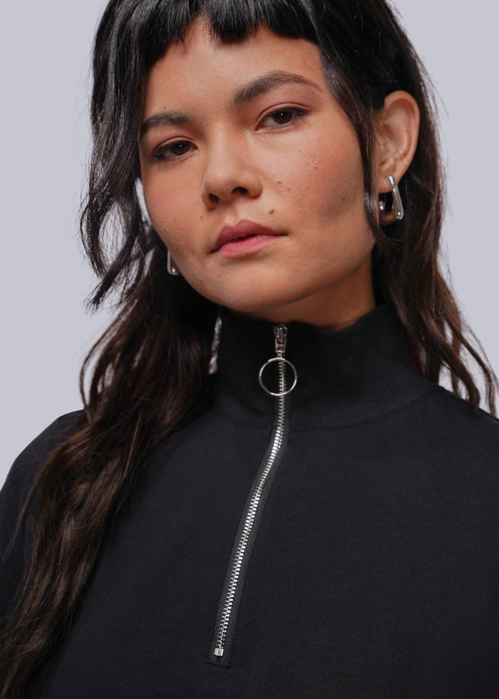 S=A close-up of a model wearing a black mock neck top with a silver zipper and a circular metal ring detail at the collar, paired with silver hoop earrings, against a solid gray background.