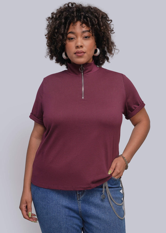 1X=A woman modeling a short-sleeve mock neck top in burgundy with a silver zipper, paired with blue jeans, against a solid gray studio background.