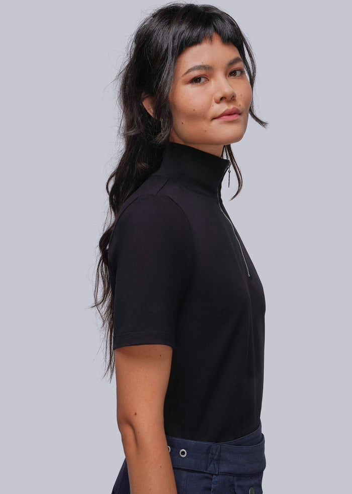 S=A woman with dark hair wearing a black short-sleeve mock neck top with a half-zip collar, paired with dark blue jeans, shown from a side profile against a solid gray background.