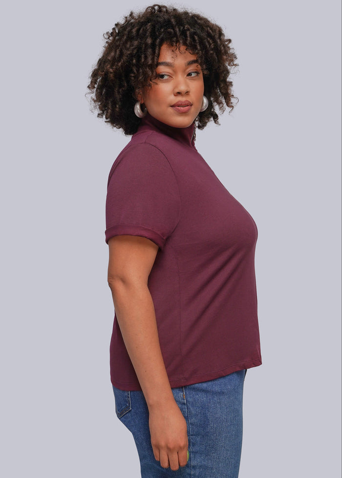 1X=A woman with curly dark hair is shown in profile wearing a short-sleeve mock neck t-shirt in a deep plum purple color, paired with blue jeans, against a solid light gray background.