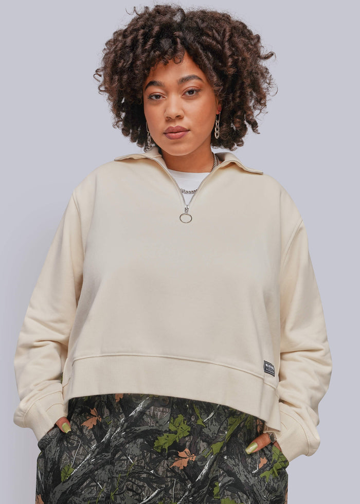 1X=A model with curly dark hair wearing a cream-colored quarter-zip sweatshirt and camouflage print pants, standing against a solid light gray background.