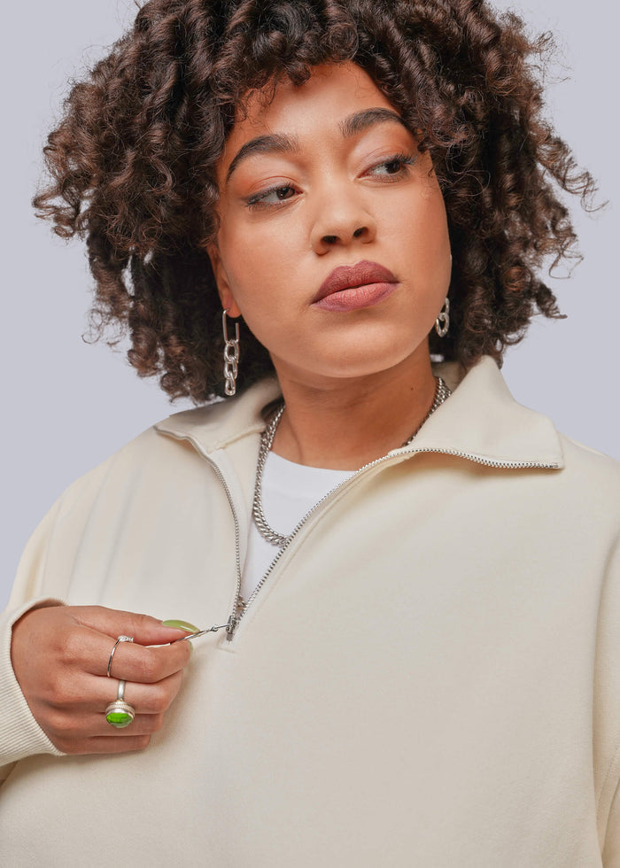 1X=A close-up of a person with curly hair wearing a cream-colored quarter-zip pullover sweater over a white t-shirt, accessorized with silver jewelry, against a solid light gray background.