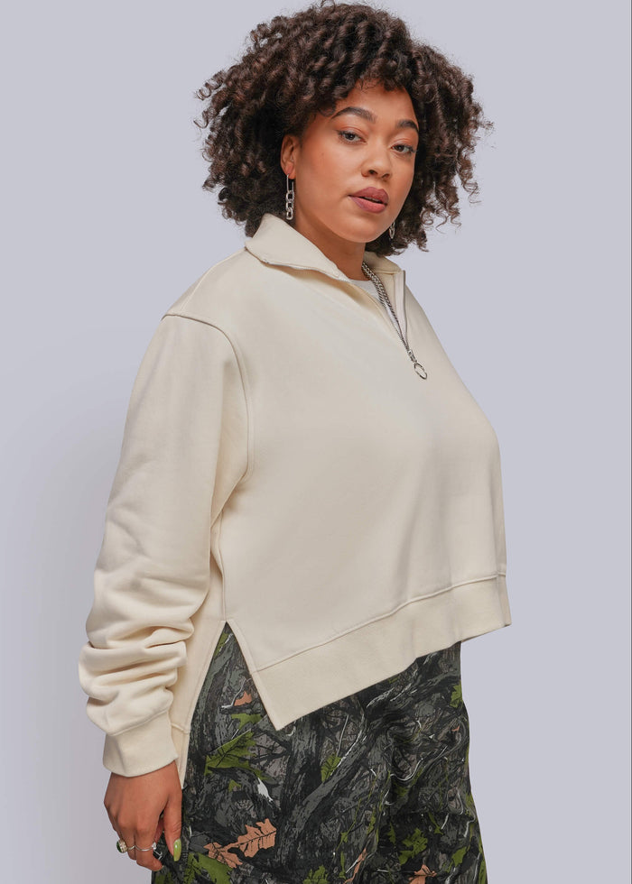 1X=A woman wearing a cream-colored cropped half-zip sweatshirt and dark green camouflage pants, photographed against a solid light gray background.
