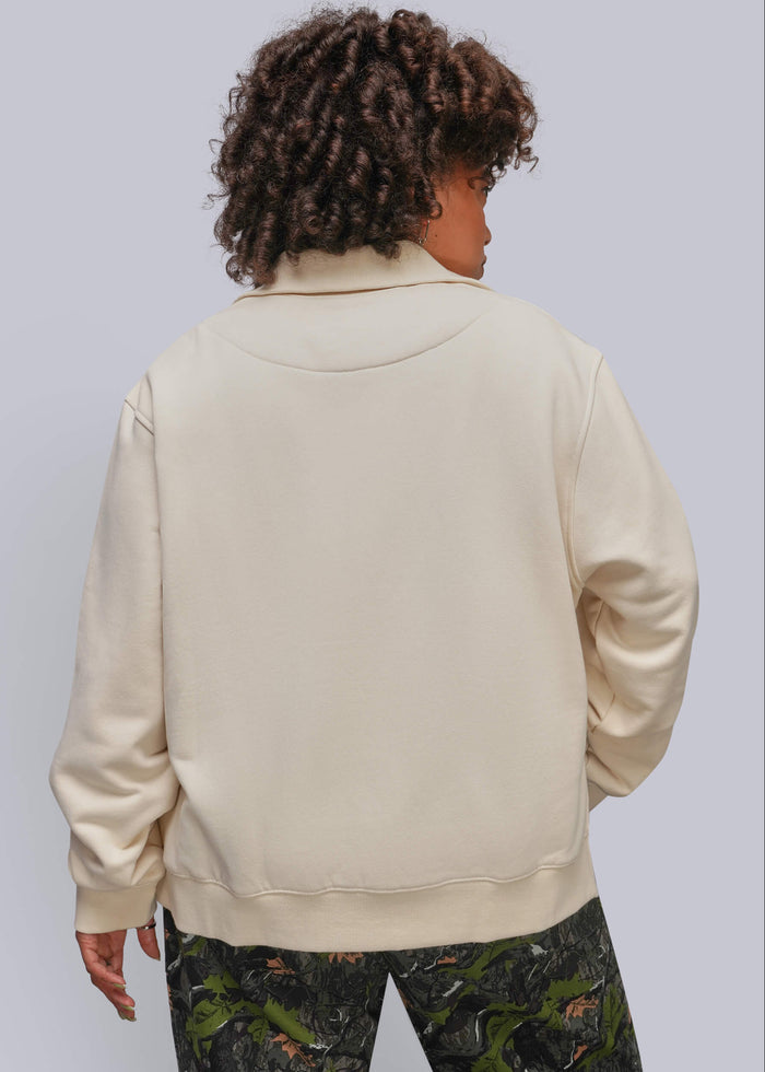 1X=A person shown from the back wearing a cream-colored oversized sweatshirt and dark camouflage pants against a light gray studio background.
