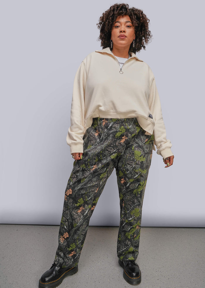 1X=A model stands against a light gray studio background wearing a cream-colored half-zip sweatshirt and dark green camouflage pants with black leather boots.