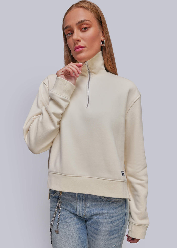 S=A woman modeling a cream-colored quarter-zip sweatshirt with a mock neck, paired with light-wash blue jeans, against a solid light gray studio background.