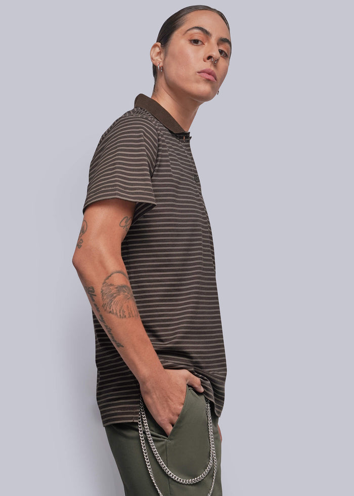 S=A person wearing a dark brown and black horizontally striped short-sleeve polo shirt with olive green pants, standing against a solid light gray background.