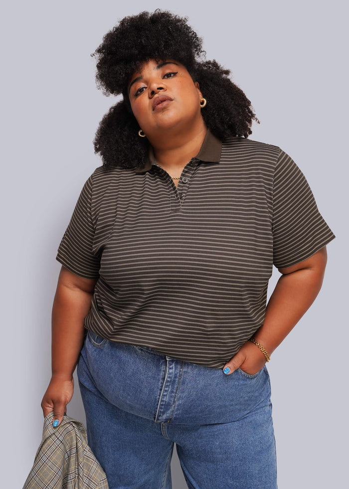 1X=A plus-size model wearing a dark brown and black horizontally striped short-sleeve polo shirt and blue jeans, holding a plaid shirt in one hand against a solid gray studio background.