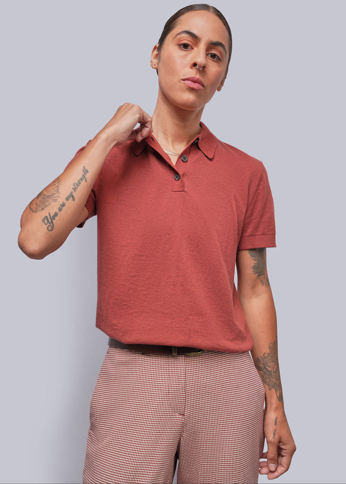 S=A model wearing a short-sleeve polo shirt in a muted terracotta or dusty rose-red color, paired with red and white houndstooth trousers, against a solid light gray background.