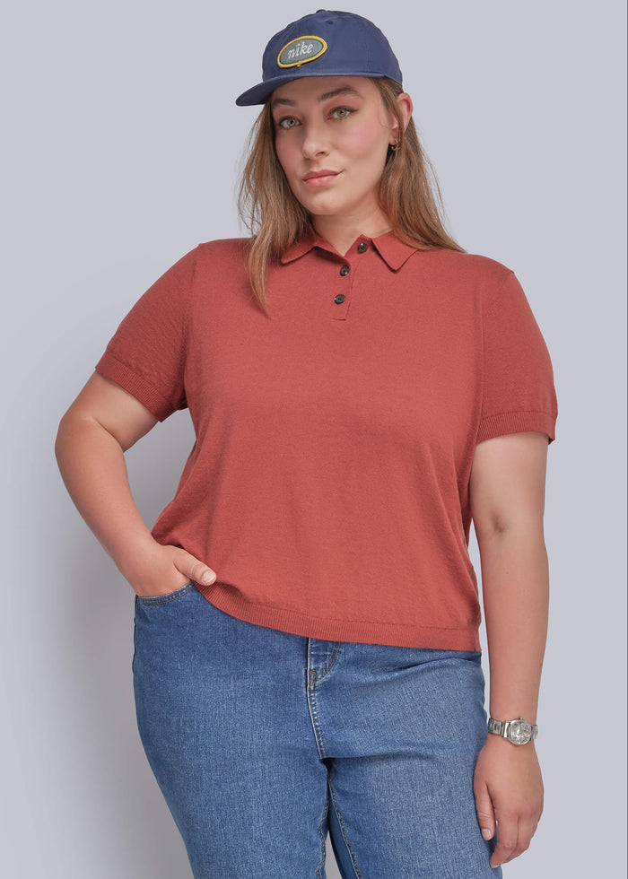 1X=A woman modeling a short-sleeve polo shirt in a muted terracotta or rust color, paired with blue jeans and a navy blue baseball cap, against a solid light gray background.