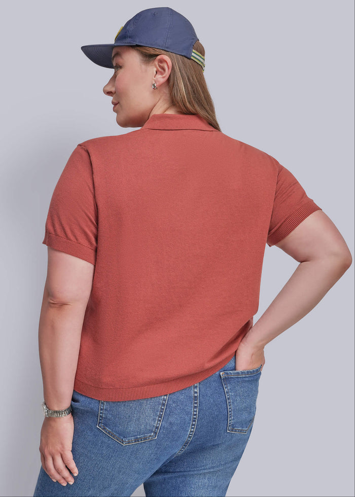1X=A woman wearing a muted terracotta short-sleeve polo shirt, blue jeans, and a navy blue baseball cap, shown from a back-three-quarter angle against a solid light gray studio background.