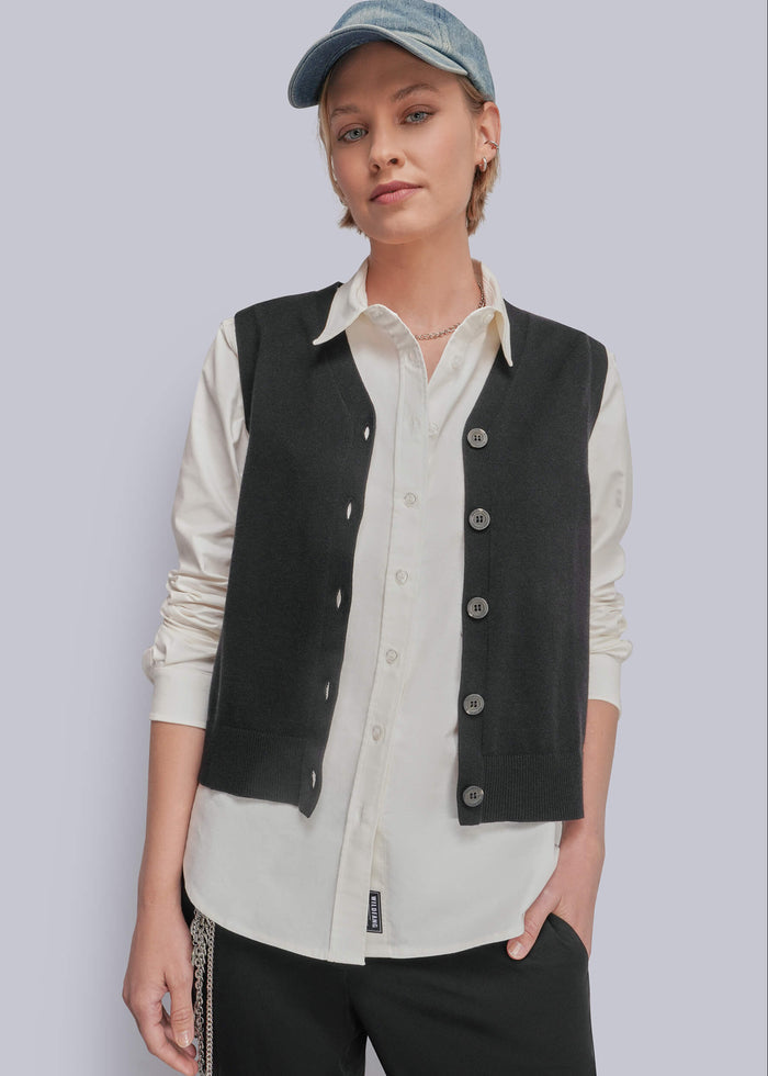 S=A model wearing a black button-up vest over a cream button-up shirt, paired with black pants and a blue denim cap, against a solid light gray background.