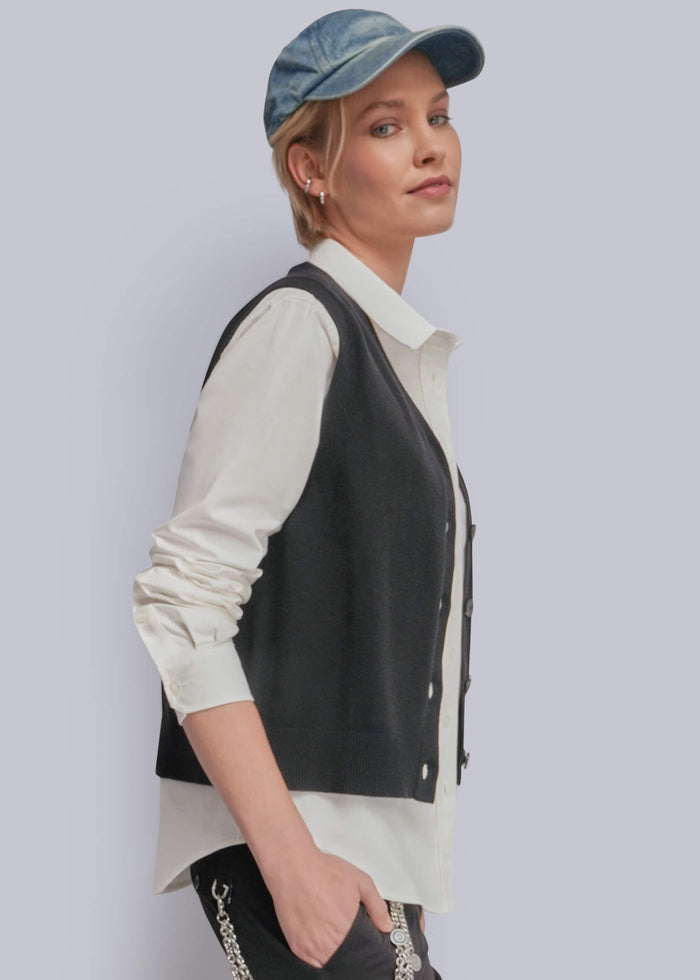 S=A woman wearing a black knit vest over a cream button-up shirt, paired with a blue denim cap and dark pants, photographed against a light gray studio background.