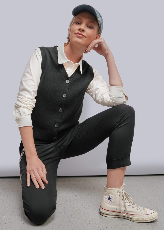 S=A model kneels on a gray floor against a light gray wall, wearing a black vest over a cream shirt, black cropped pants, and a black cap. She is wearing off-white high-top sneakers with a Converse star logo visible on the side.
