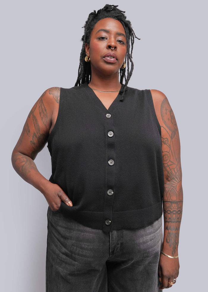 1X=A woman modeling a sleeveless black knit vest with a button-down front, paired with dark gray jeans, against a solid gray studio background.