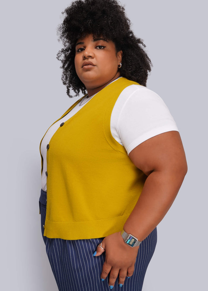 1X=A plus-size model wearing a mustard yellow sleeveless knit vest over a white short-sleeve top and navy blue pinstriped trousers, photographed against a solid light gray background.