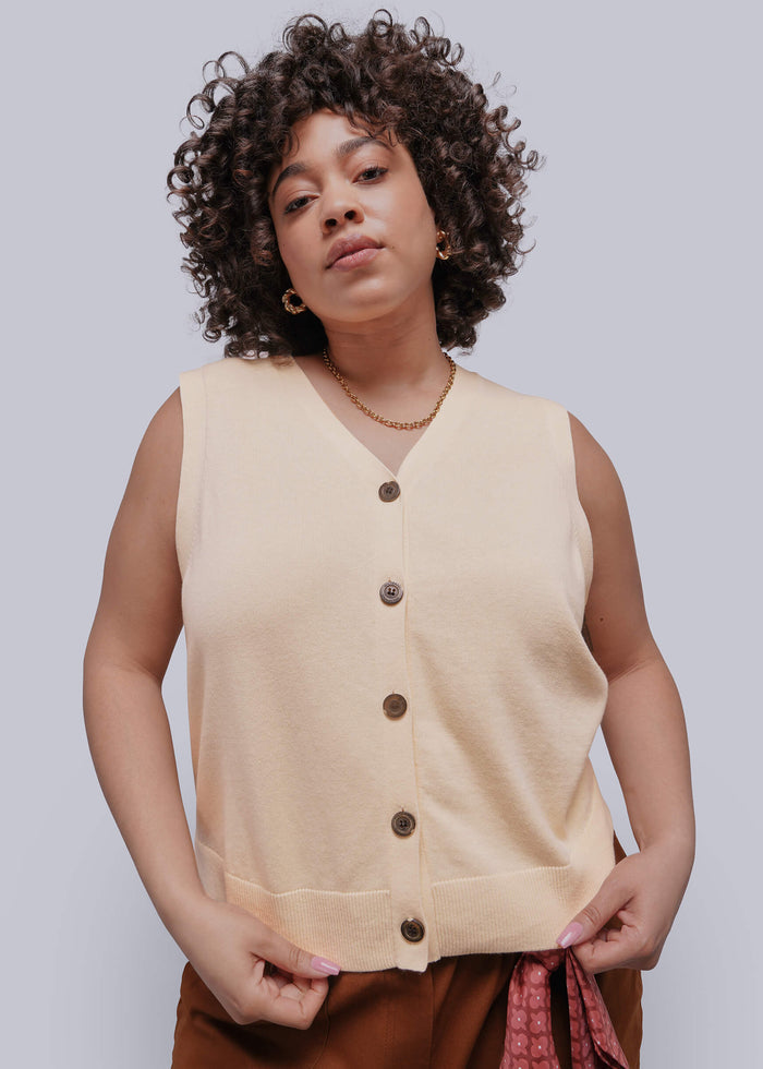 1X=Woman wearing a beige sleeveless sweater vest with black buttons against a plain background