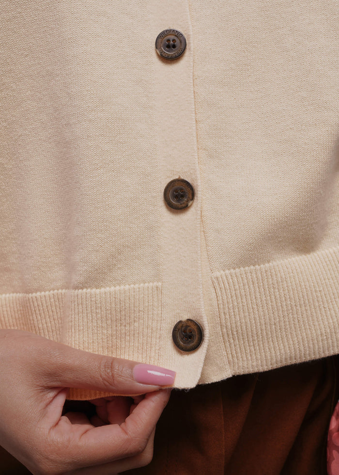 1X=Close-up of a beige sweater vest with buttons and a hand adjusting it.