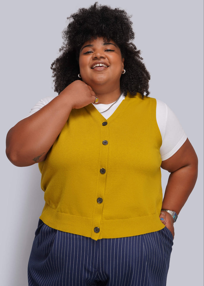 1X=A plus-size model wearing a mustard yellow button-up knit vest over a white t-shirt and navy blue pinstripe trousers, posing against a solid gray studio background.