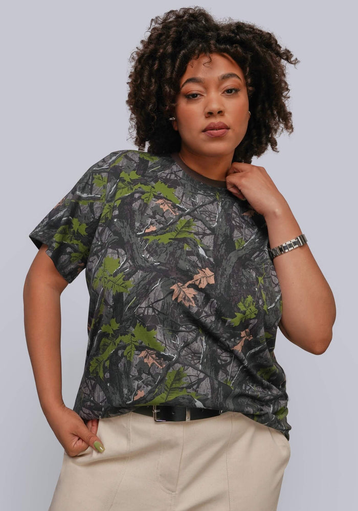 1X=A woman modeling a short-sleeve crewneck t-shirt with a dark gray and green woodland camouflage pattern, paired with beige pants, against a solid light gray background.