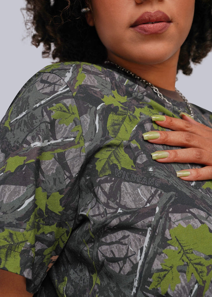 1X=Close-up of a person wearing a camouflage short-sleeve top with a woodland tree-bark and leaf pattern in gray, green, and black. The word 'WILDFANG' is visible on the chest.